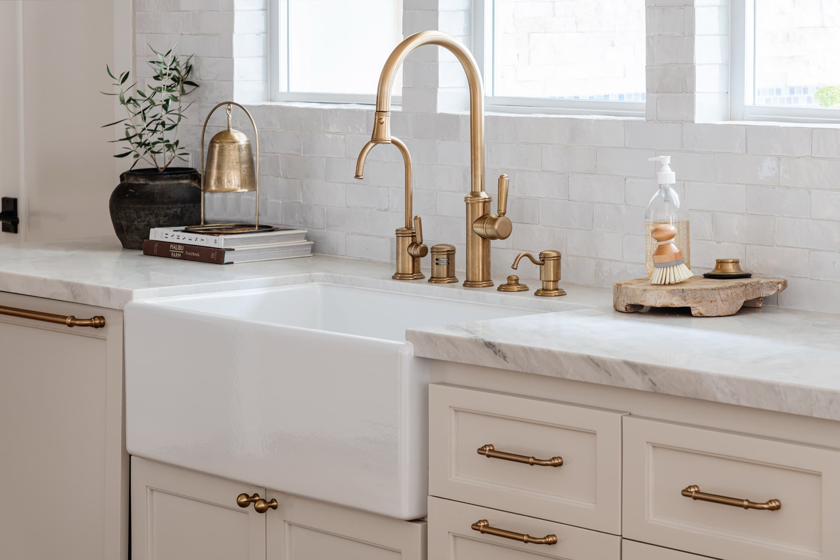 An apron-front farmhouse sink under a window, with an aged-brass tap and stoneware on the counter.