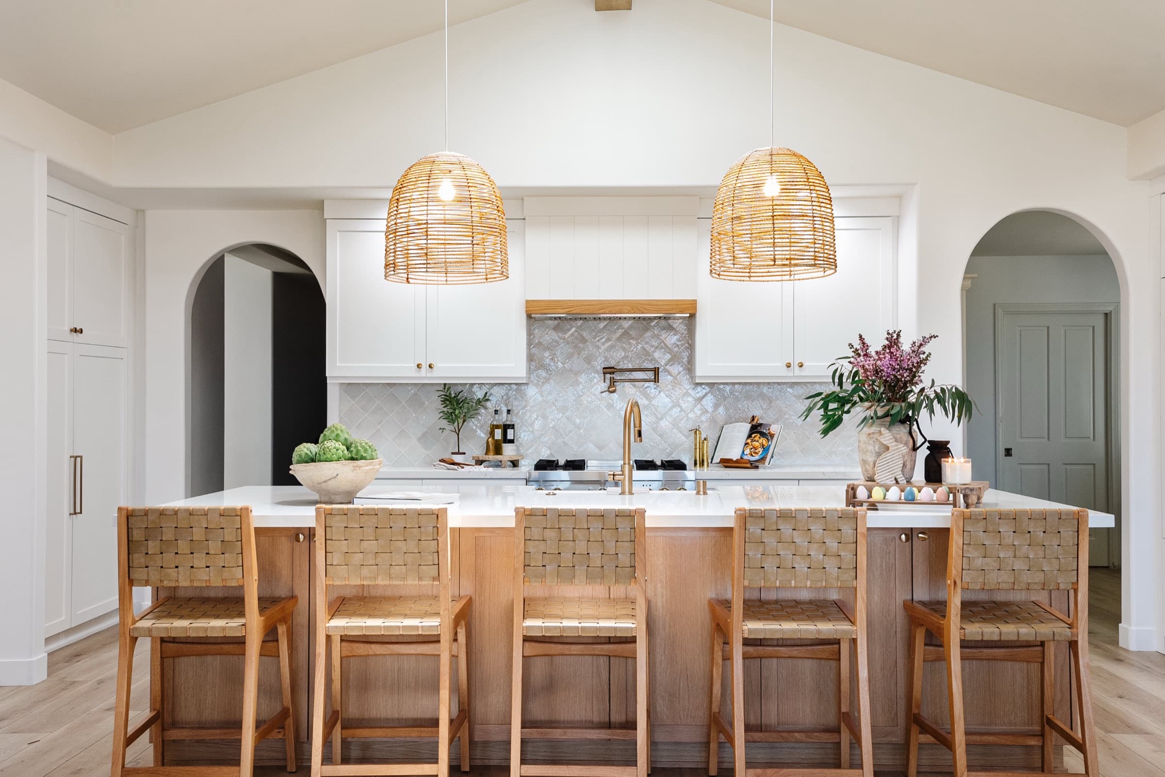 A vaulted white kitchen with two woven pendants over a long white-oak island and five leather-strap counter stools.