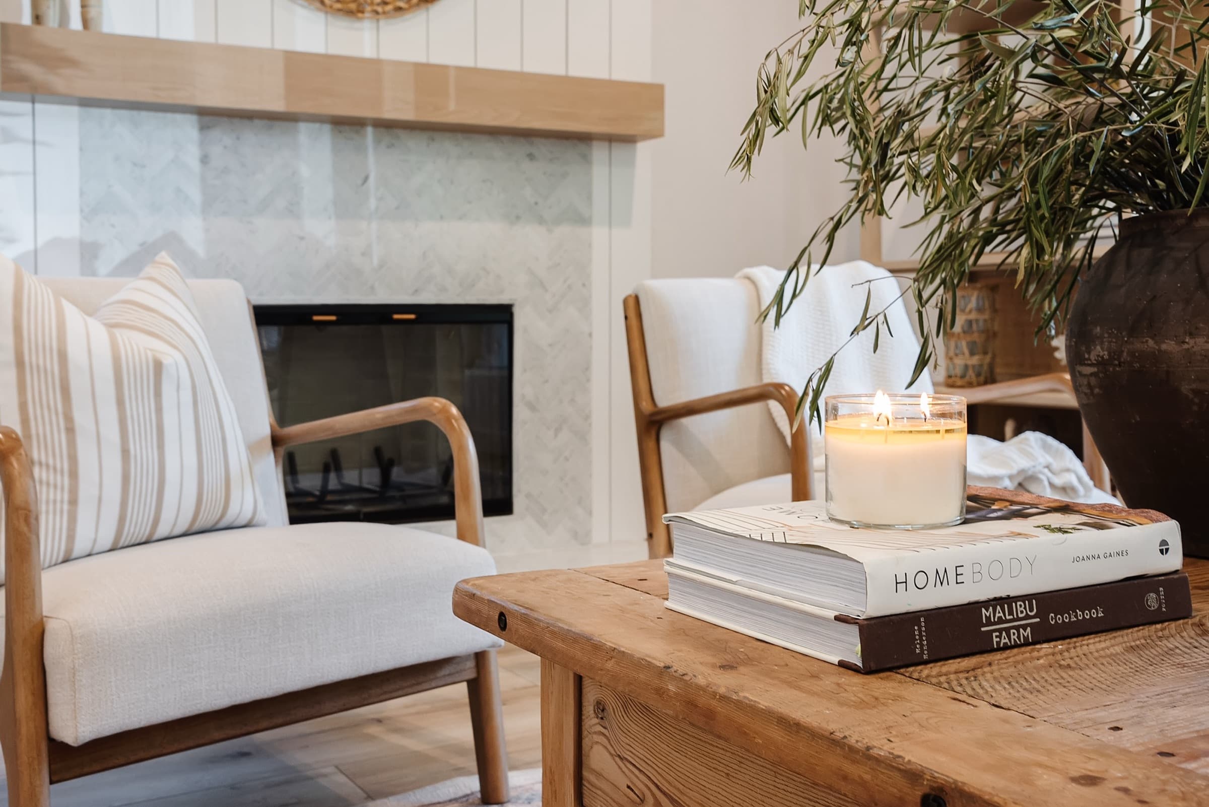 A lit candle and a stack of books on a reclaimed-wood table, with two linen armchairs and a marble fireplace behind.